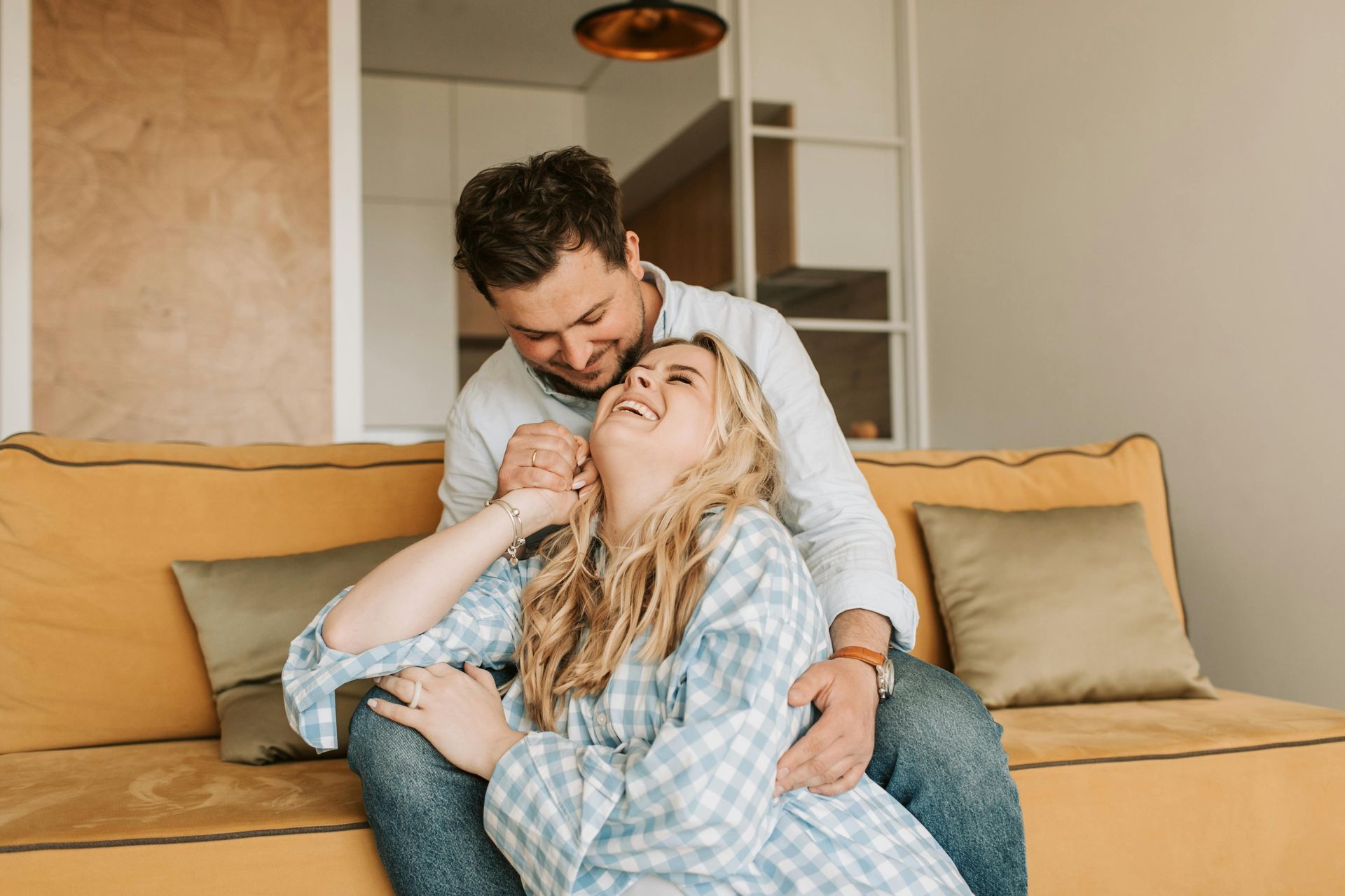 Couple cuddling on a tan sofa, smiling in a cozy living room.