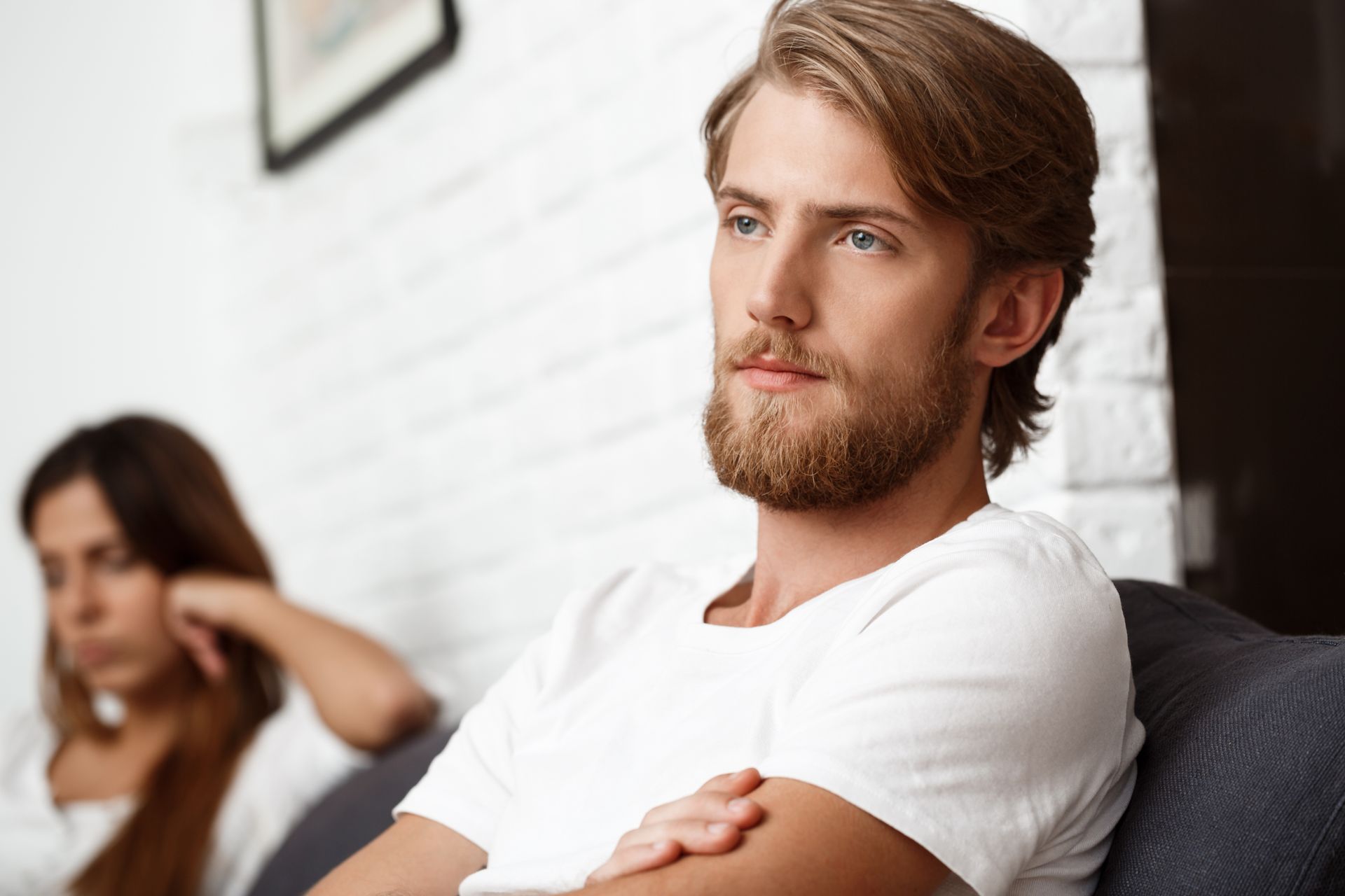 Man sitting with arms crossed on a couch, looking away; blurred woman in the background.