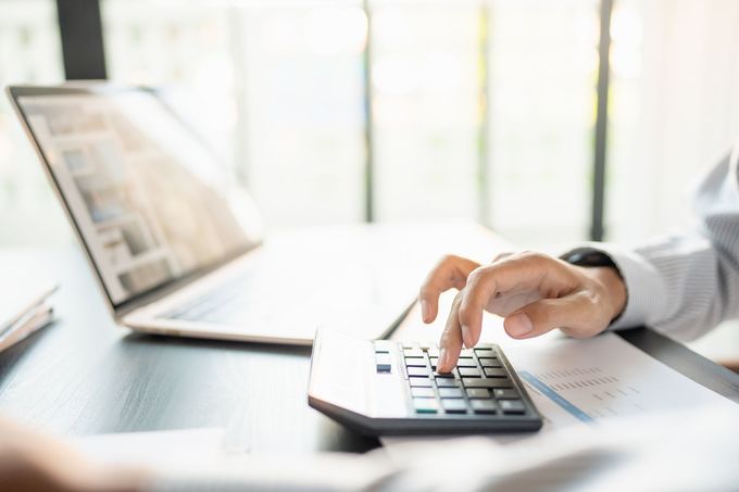 Hand using a calculator beside a laptop on a bright office desk