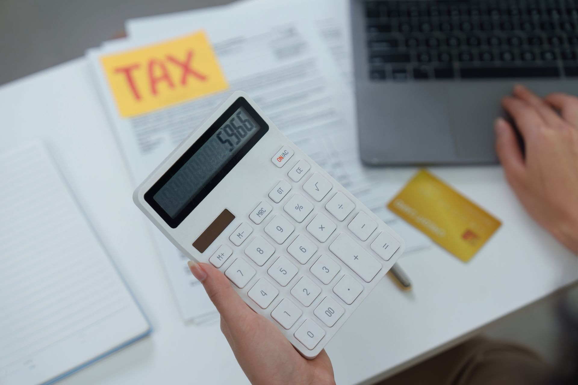 Hand holding calculator over tax forms beside laptop and credit card on desk