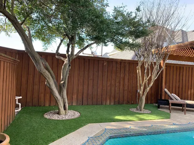 A backyard with a pool, trees, and a brown fence. Green artificial grass and a pergola are also visible.
