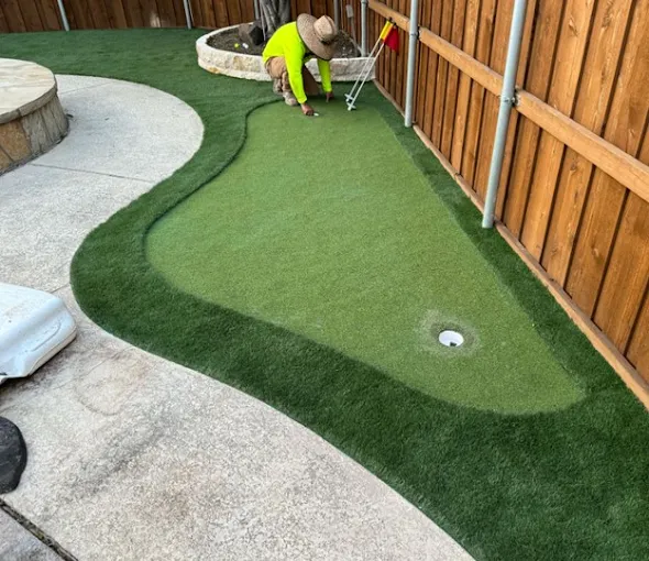 Person installing flag on a backyard putting green with a concrete path and wooden fence.