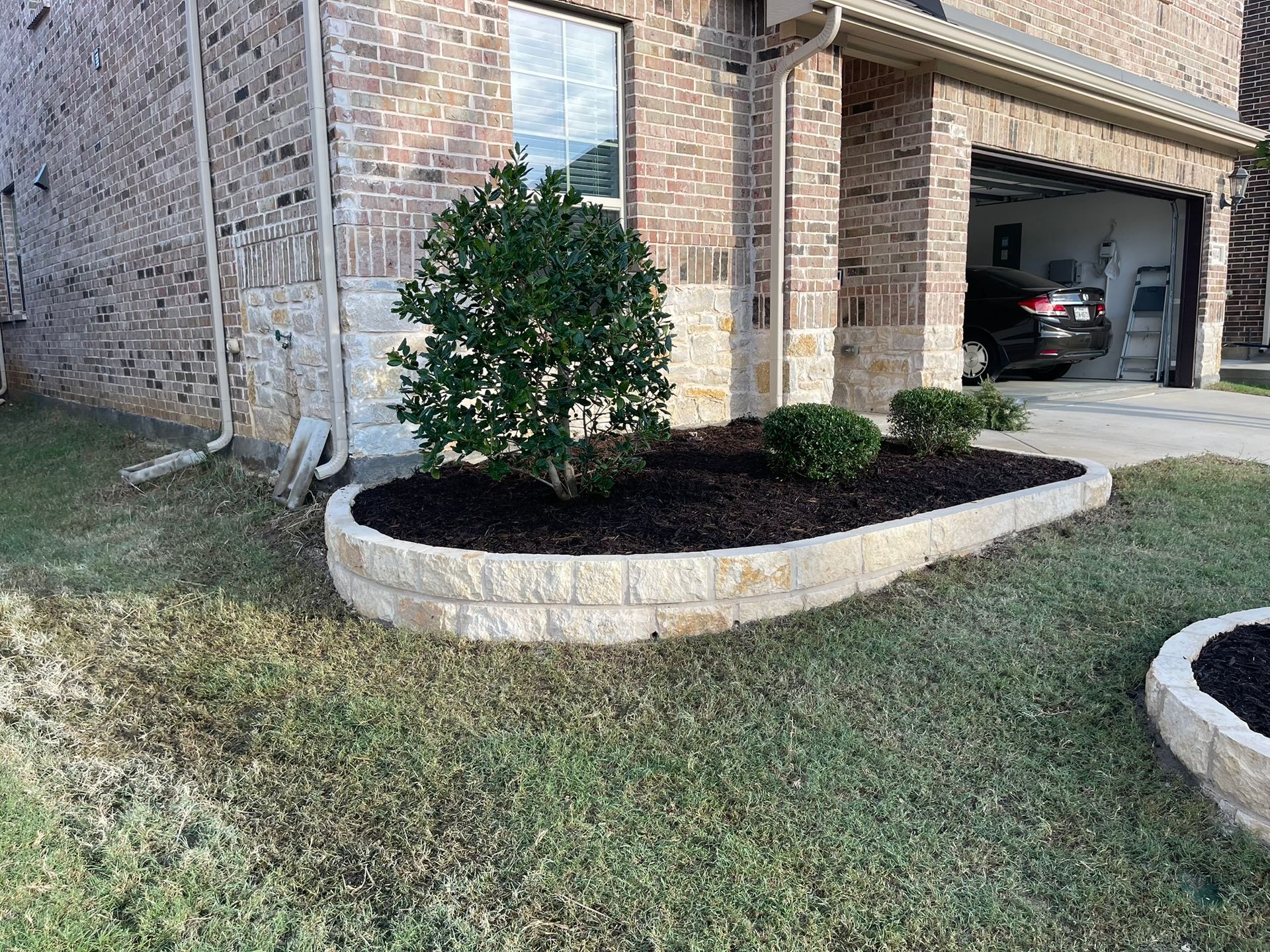 Raised stone flower bed with green plants, brown mulch, and brick house.