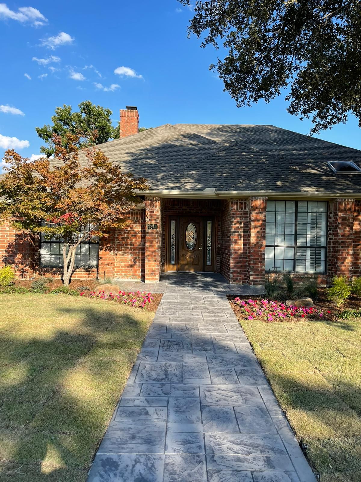 Brick house with gray walkway, flowers, green grass, and a clear blue sky.