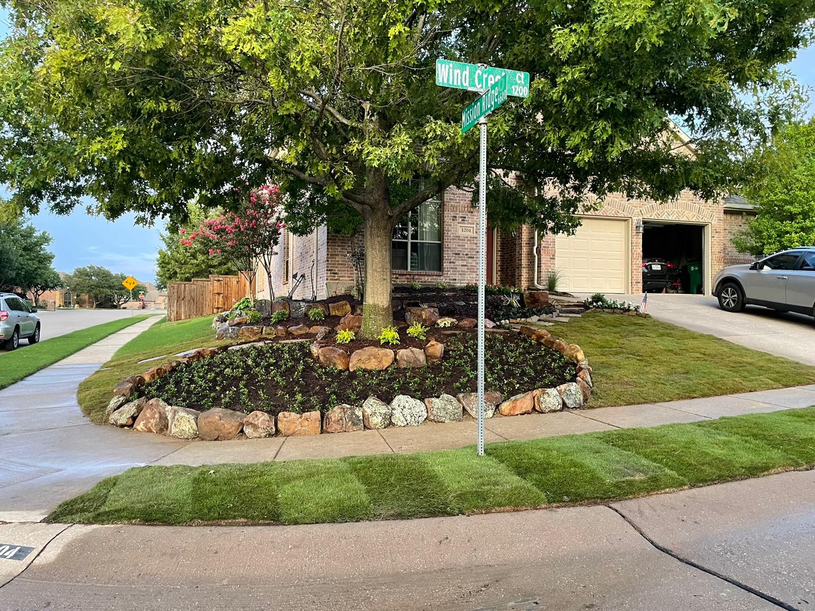Corner lot with a tree, landscaped bed, and street sign in front of a brick house.