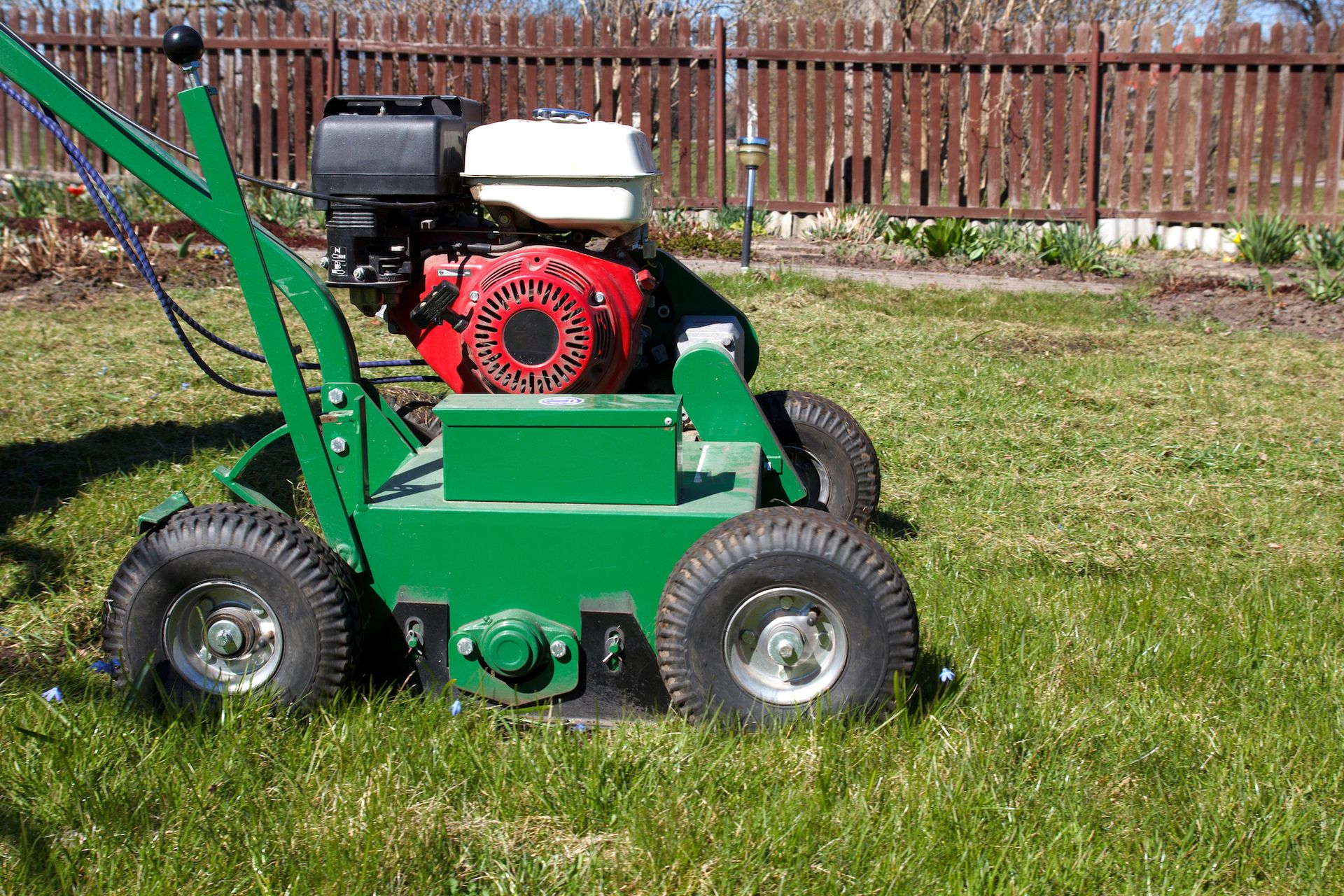 Green lawn dethatcher with a red engine, operating on a green lawn near a fence.