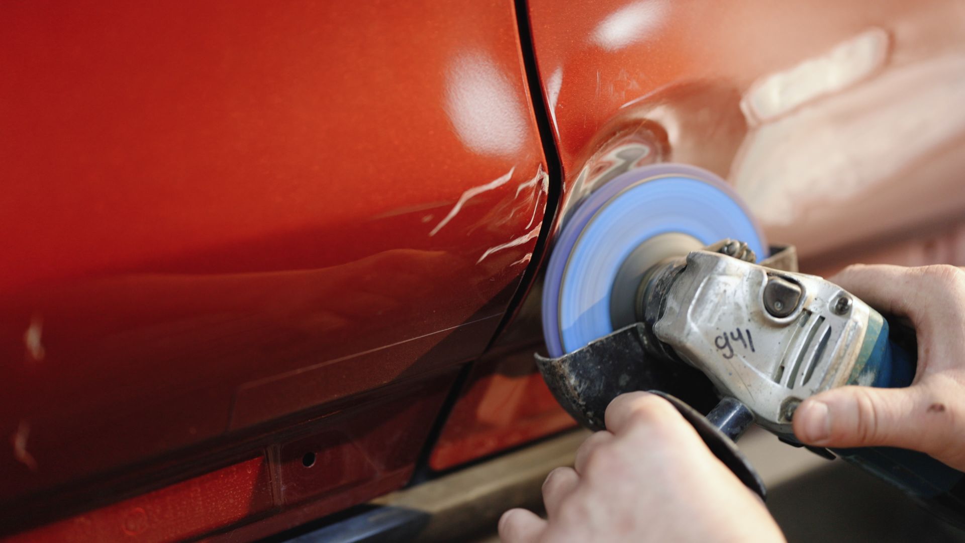 Hands using a power grinder to smooth damage on a red car panel in an auto body shop. Hands using a power grinder to smooth damage on a red car panel in an auto body shop.