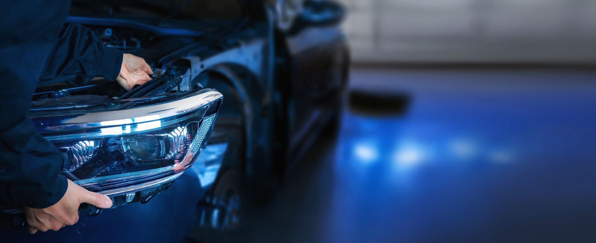 Hands removing a car headlight from the front of a vehicle in a garage.