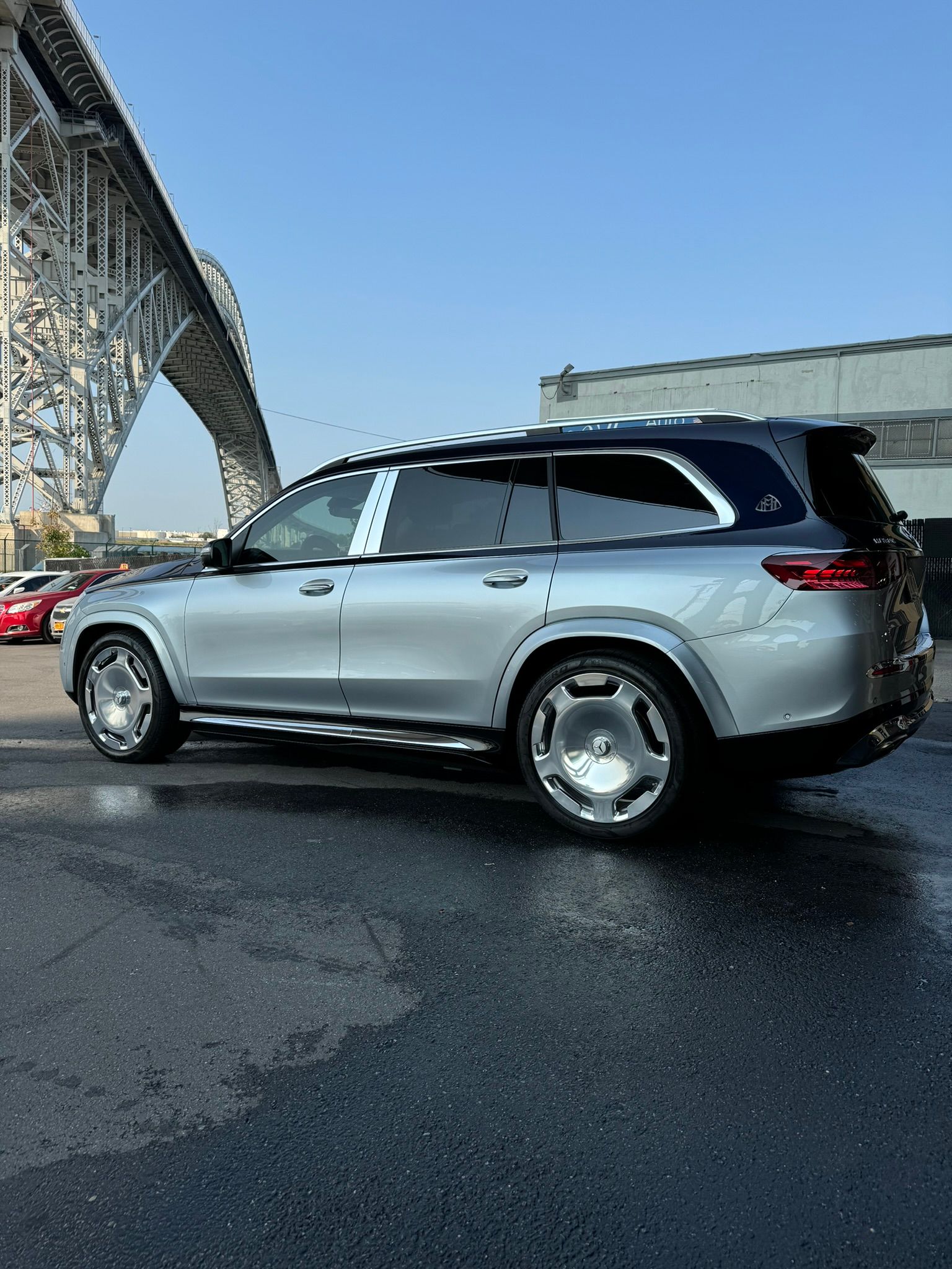 Silver Mercedes-Maybach SUV parked on wet pavement under a bridge on a sunny day.