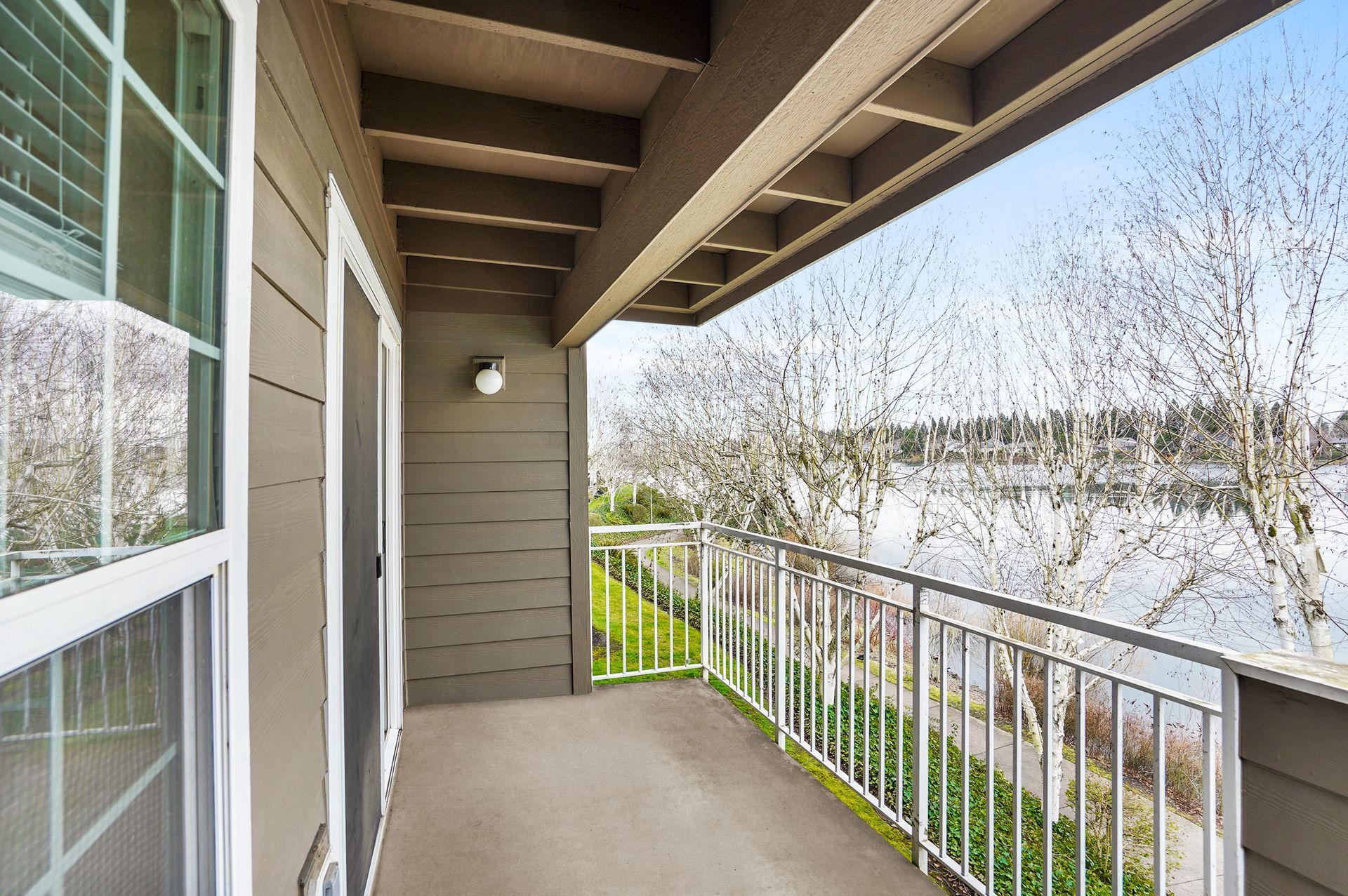 An outdoor balcony with beige walls and a white railing overlooking a lake surrounded by bare trees under a clear sky.