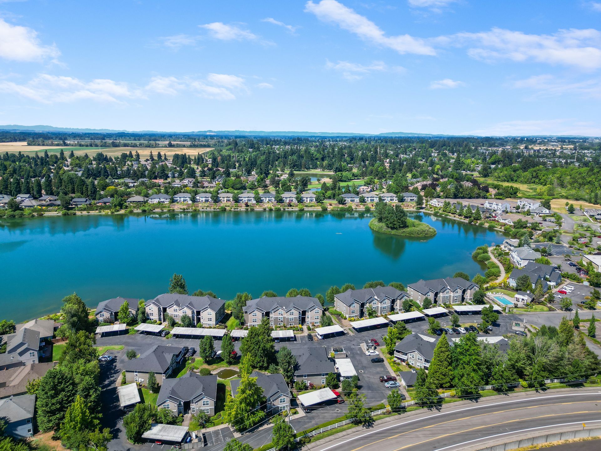 Aerial view of an apartment complex with multi-story buildings and a large blue lake, surrounded by trees and a road.