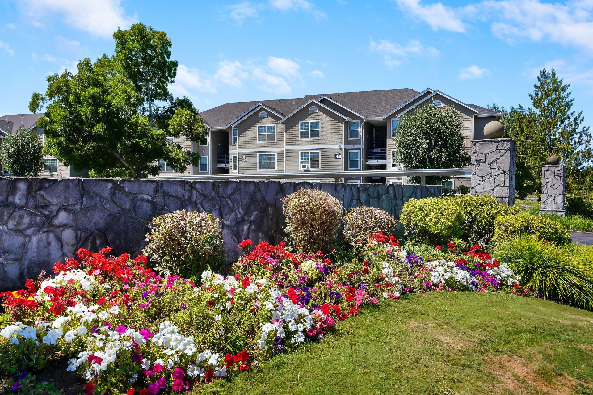 A multi-story apartment building sits behind a stone retaining wall filled with colorful flowers and green shrubbery.
