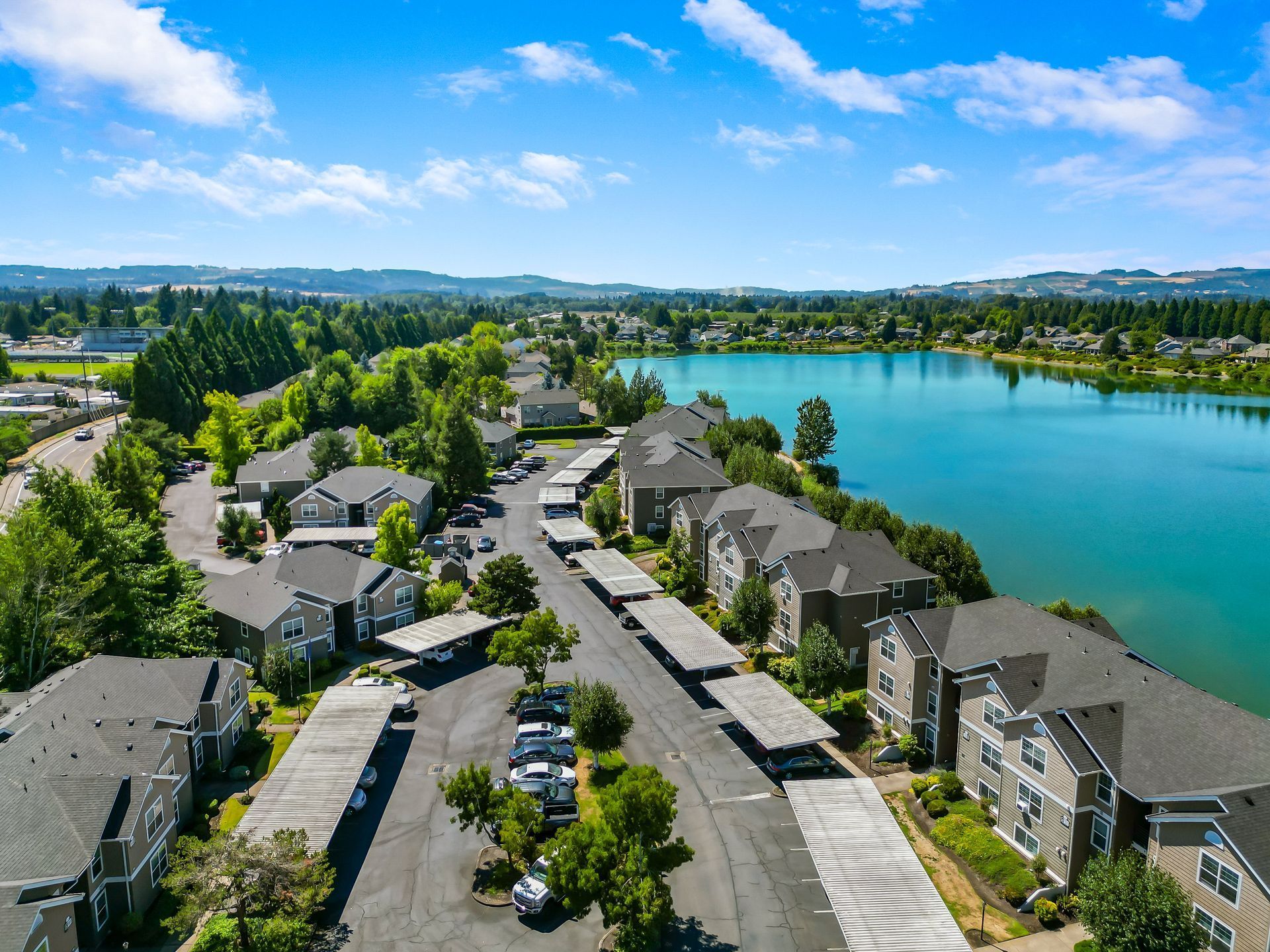 An aerial view of an apartment complex with tan buildings, paved parking with carports, and a vibrant blue lake nearby.