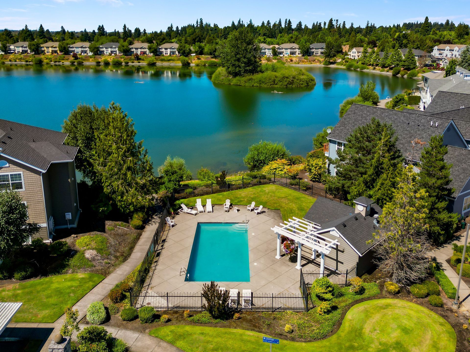 An aerial view of a rectangular outdoor swimming pool with a white pergola, set beside a lake and apartment buildings.