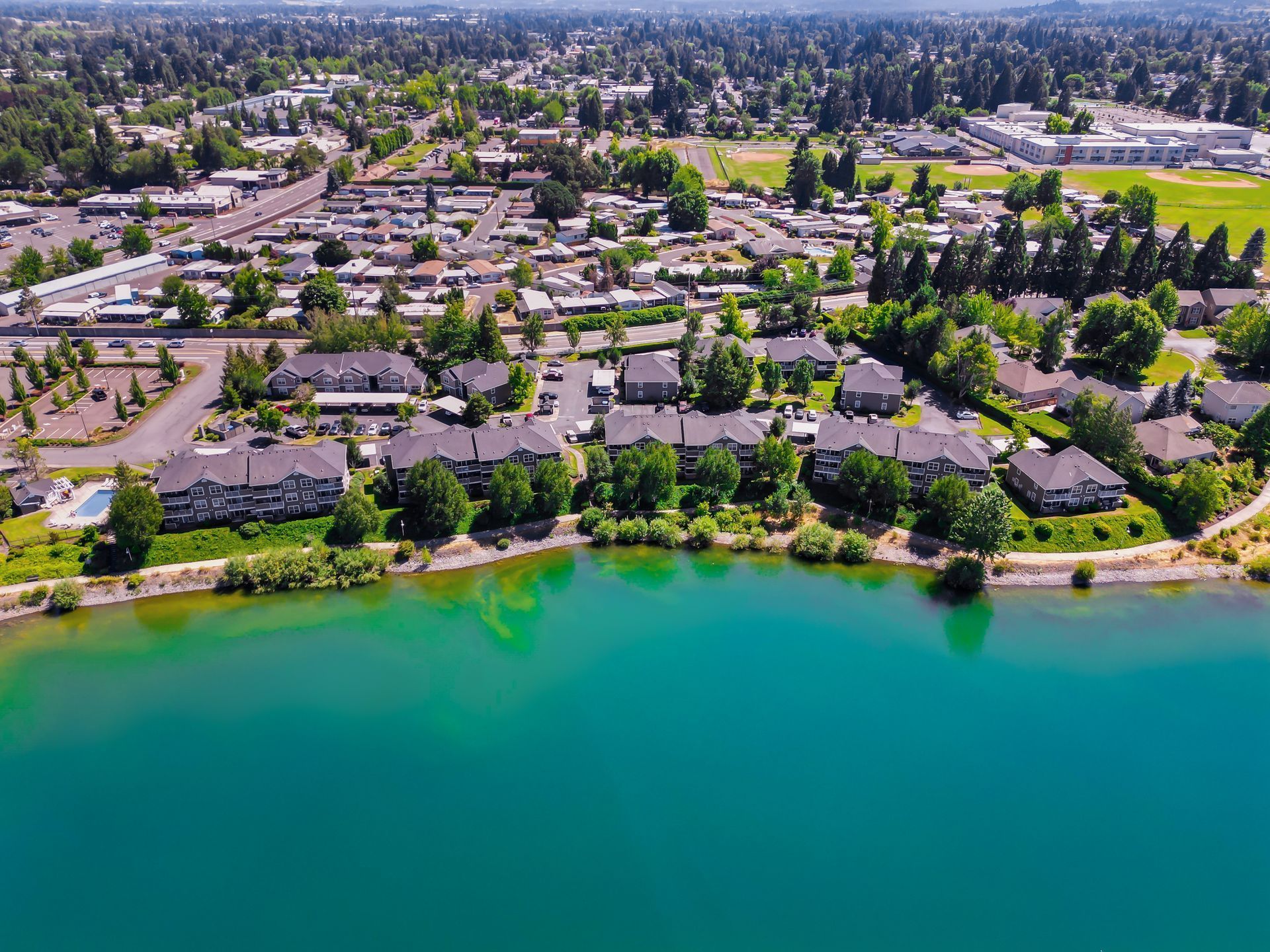 Aerial view of multi-story apartment buildings situated along the shore of a vibrant turquoise lake in a suburban area.