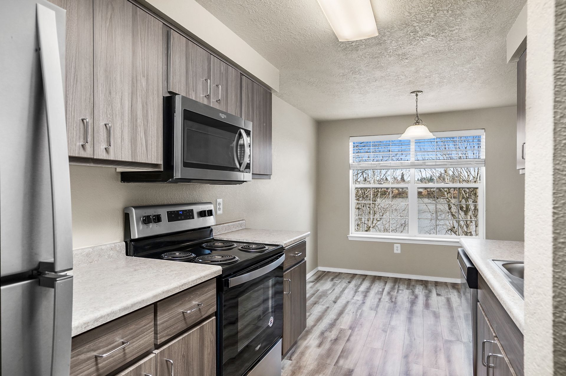 A kitchen with gray wood-look cabinets, stainless steel appliances, light countertops, and a window in the dining area.