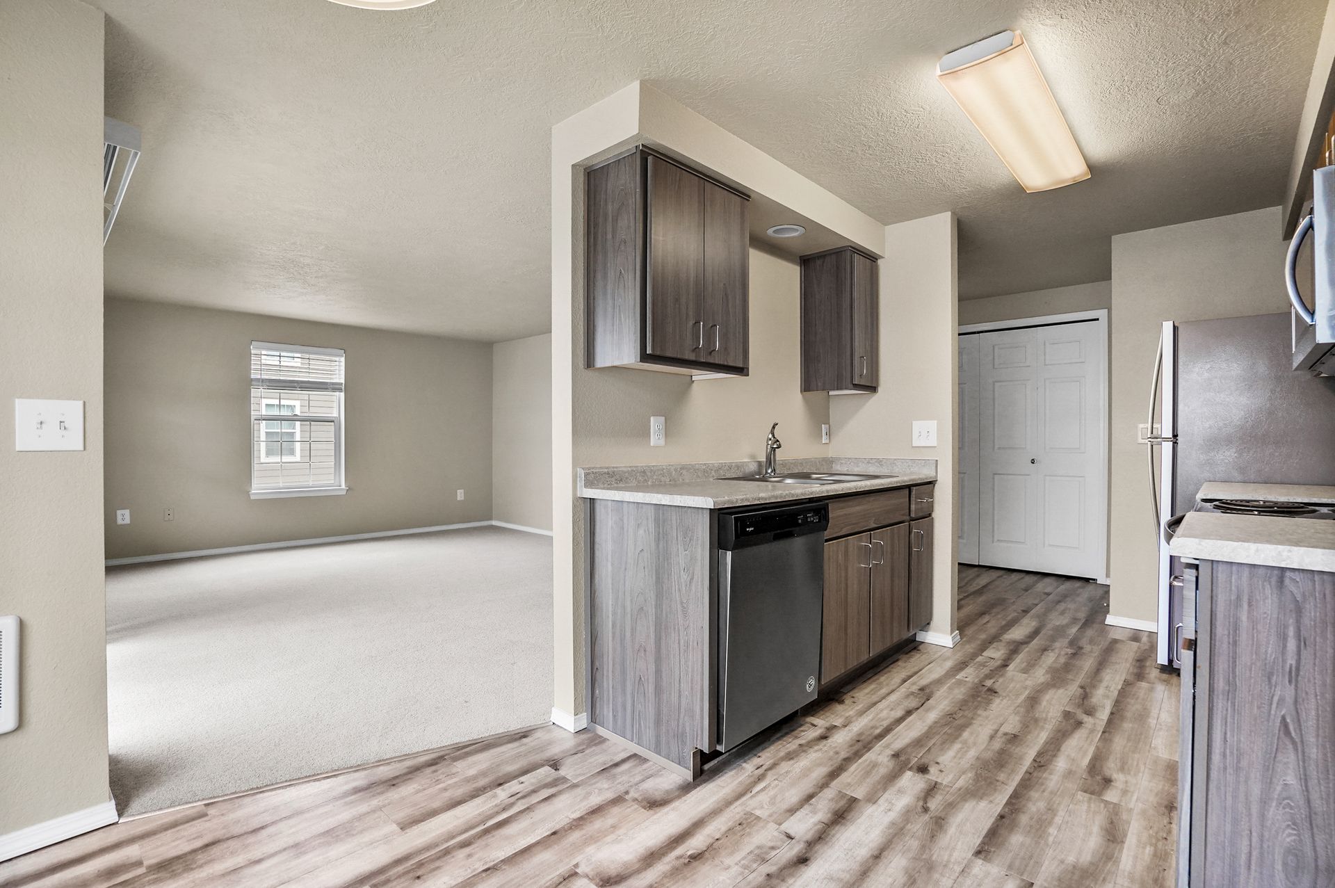 A kitchen with wood-look flooring and dark cabinets opens into an adjacent living area with carpet and a large window.