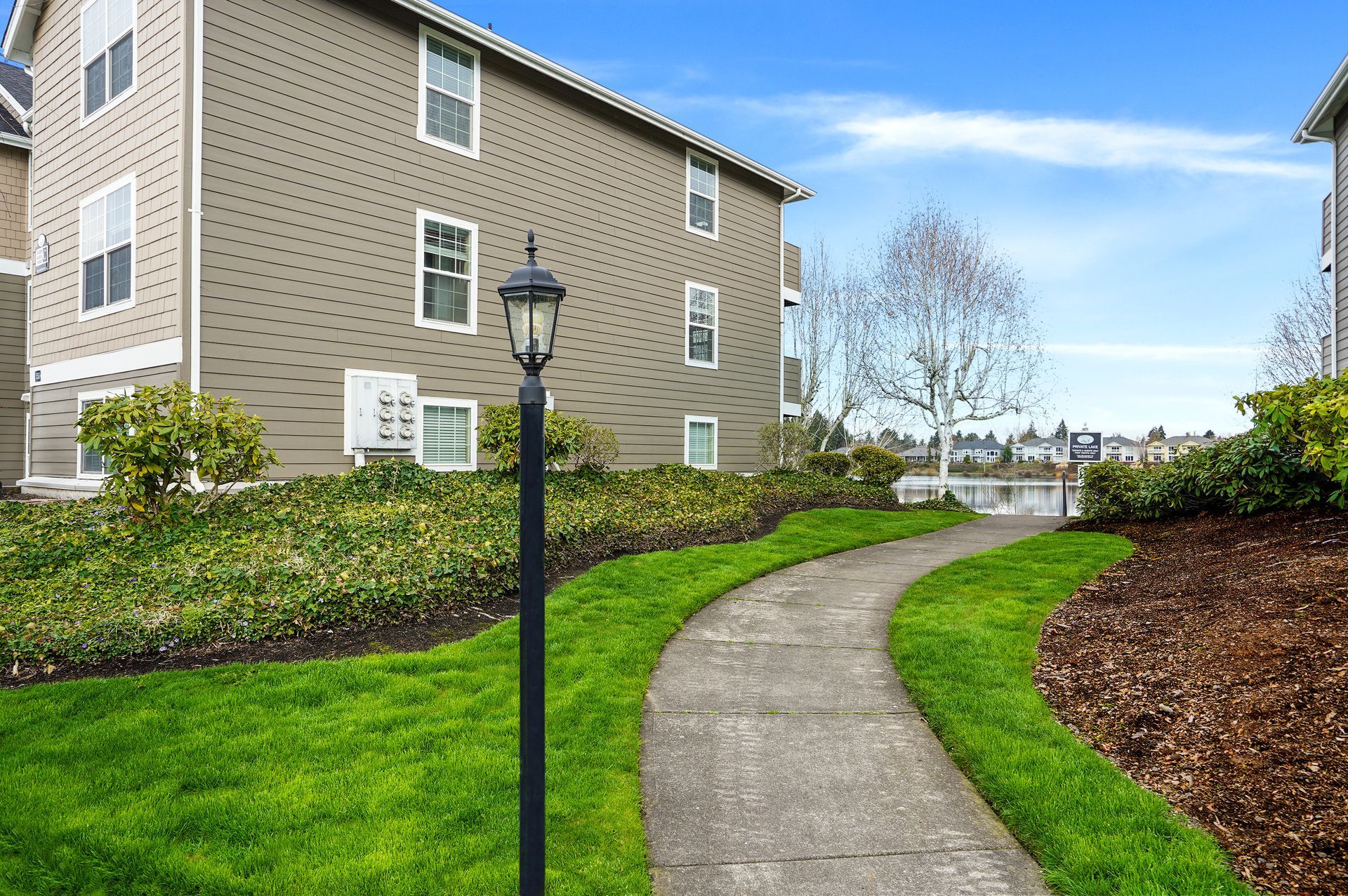 A paved walkway leads past a tan apartment building with a lamppost, green lawn, and shrubbery under a blue sky.