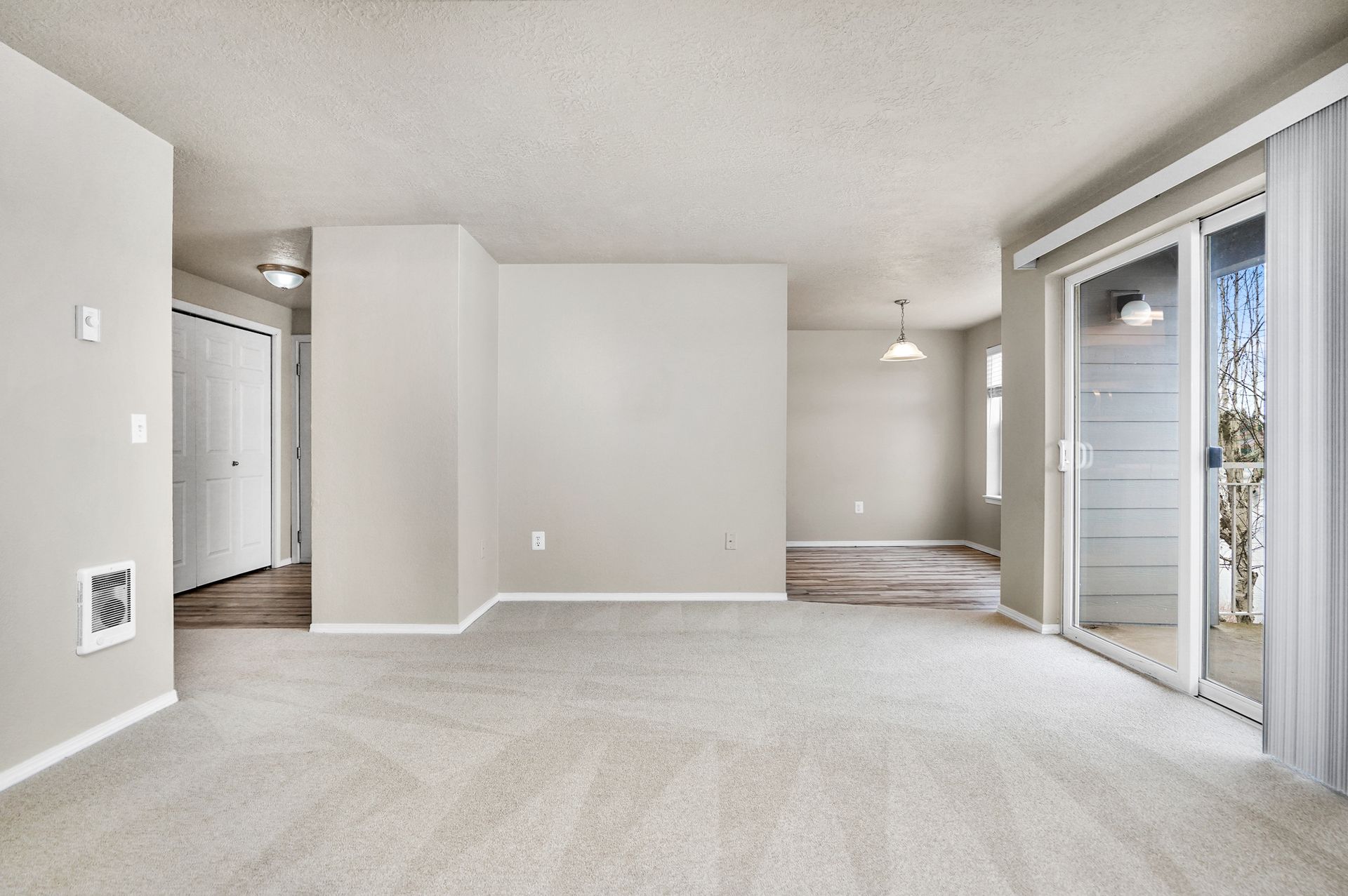 A brightly lit, empty living room with light beige walls, tan carpeting, and a large sliding glass door leading outside.