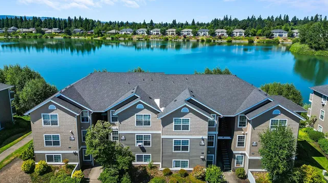 An aerial view of a multi-story apartment building with grey siding and a dark roof, situated beside a bright blue lake.