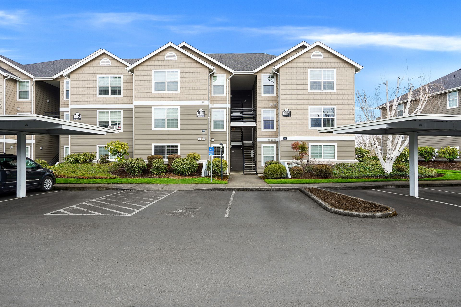 A three-story apartment complex with beige siding, covered parking, and a paved lot under a blue sky.