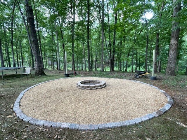 Gravel fire pit ring in a wooded park clearing, bordered by stones and surrounded by trees.