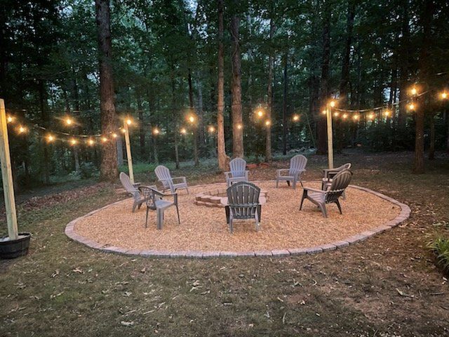 String lights over a circular gravel patio with several chairs in a wooded backyard at dusk