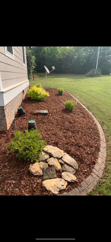 Mulched garden bed along house with small shrubs, stepping stones, and a lawn.