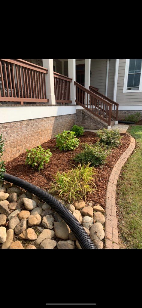 Landscaped front yard with mulch, shrubs, stones, and a porch beside a brick house.