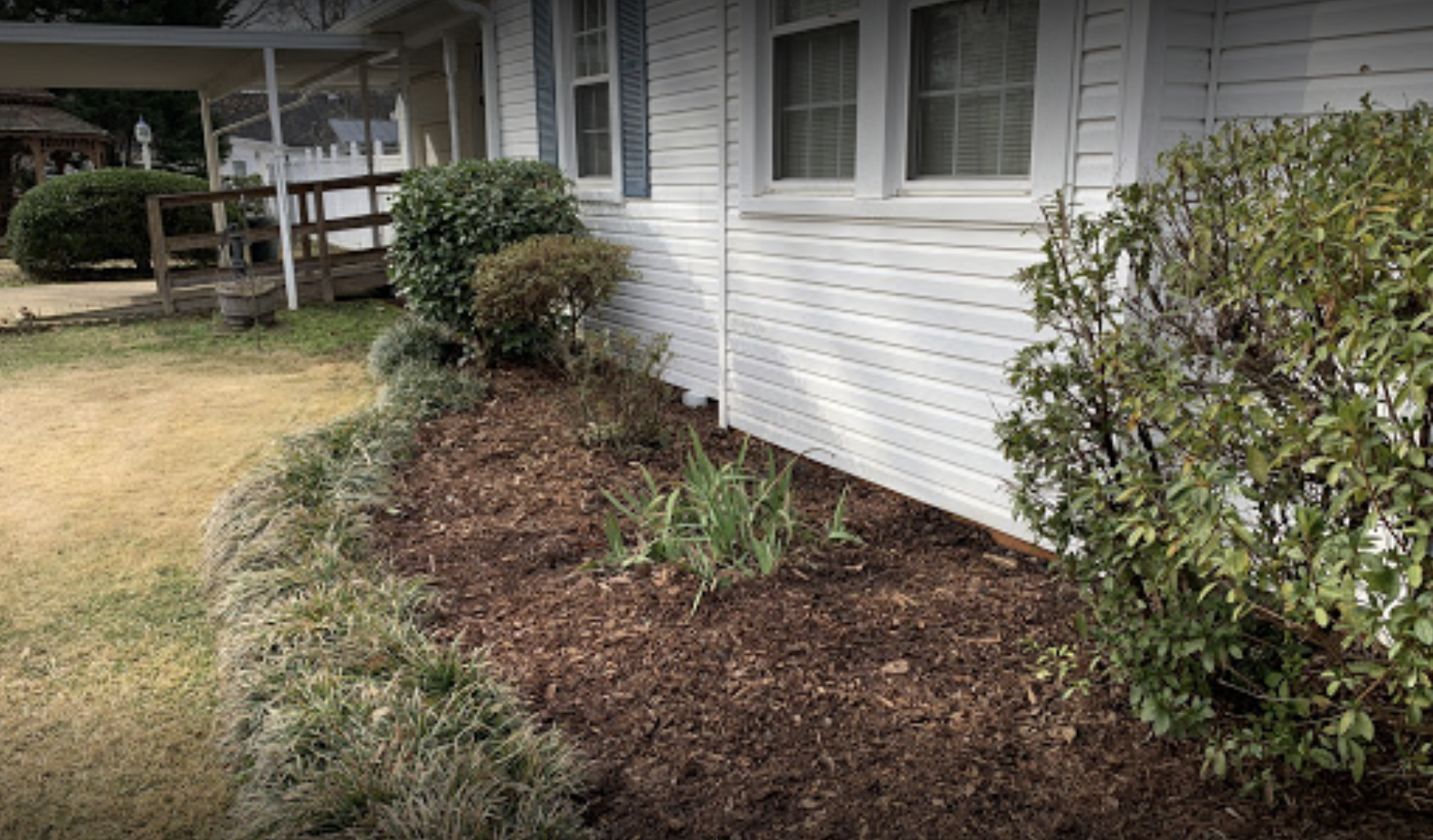 Mulched garden bed along white house exterior with shrubs and a porch in the background