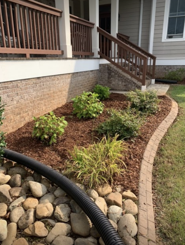 Mulched garden bed with shrubs and stone border beside a house porch and brick foundation