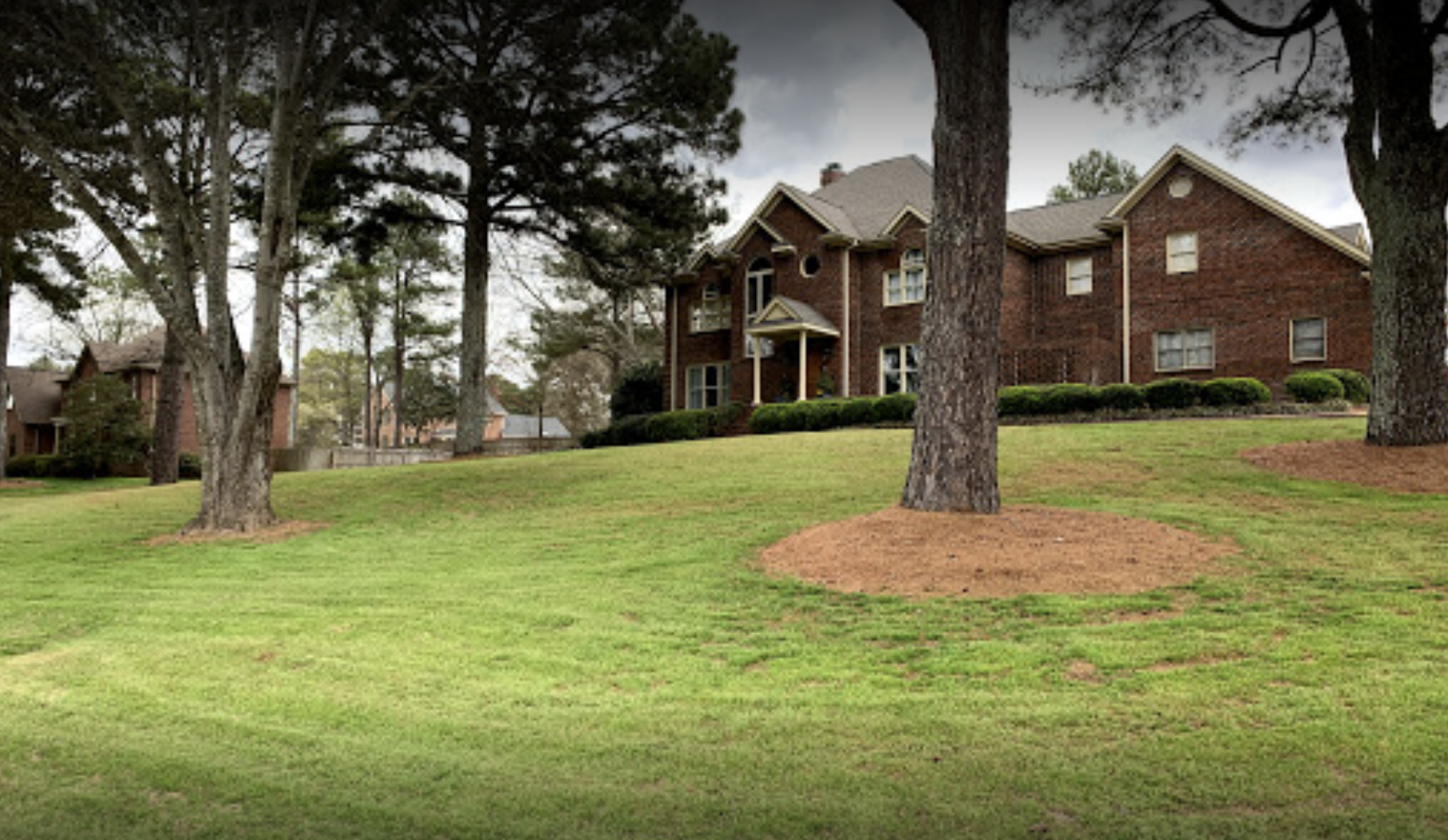 Brick apartment building behind grassy lawn with pine trees on a cloudy day