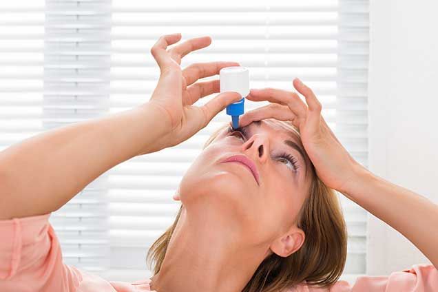 Woman applying eye drops, looking up, holding eyelids open with fingers.