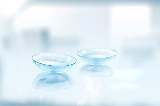 Two clear contact lenses in a light blue liquid, reflecting on a white surface.