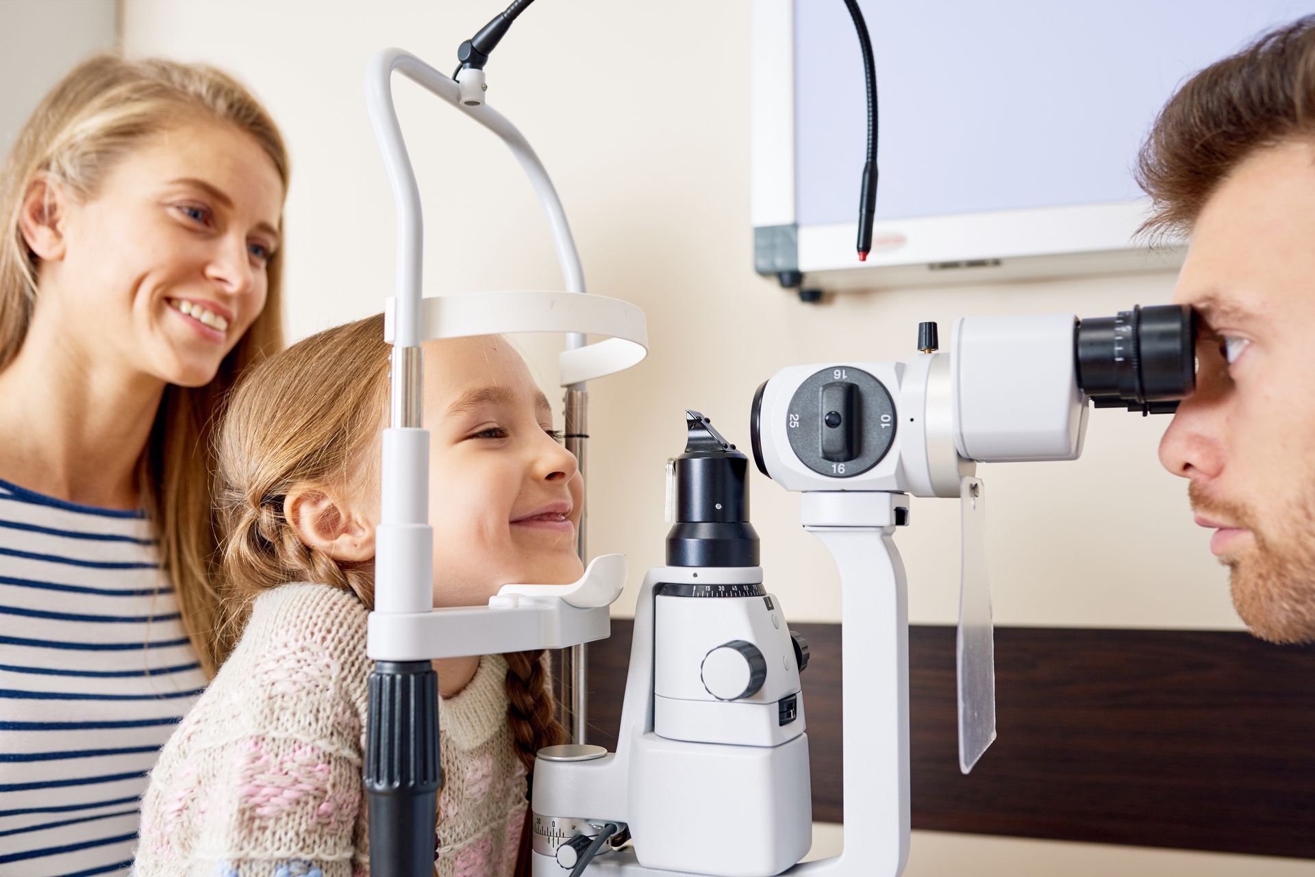 A child is receiving an eye exam using a slit lamp while an adult stands nearby. A child is receiving an eye exam using a slit lamp while an adult stands nearby.