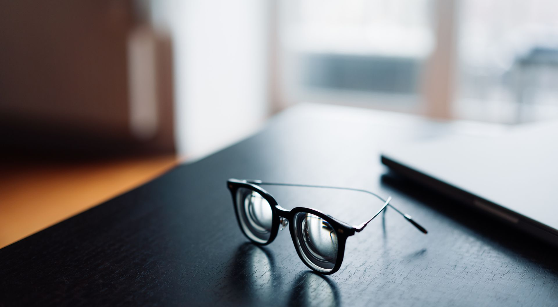 Eyeglasses on a dark table, near a blurred laptop and window.