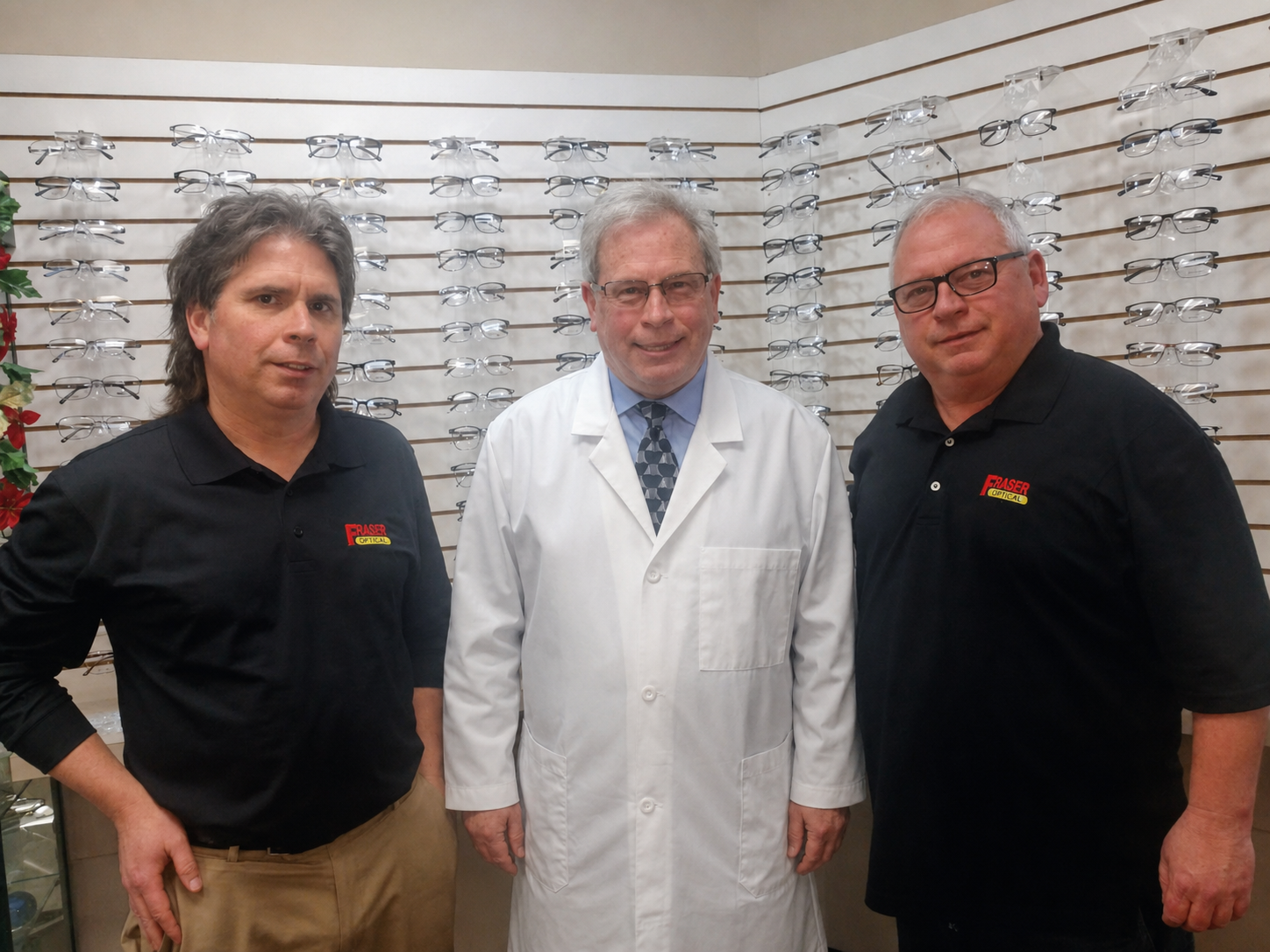 Three men standing in front of a wall of eyeglass frames, one in a white coat and two in black shirts