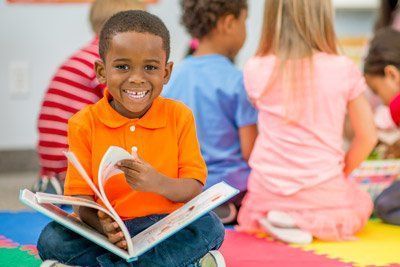 Boy smiles while reading a book in a classroom; children sit nearby.