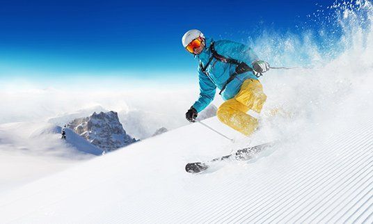 Skier carving down a snow-covered slope, blue jacket, yellow pants, blue sky, and mountains in the background.
