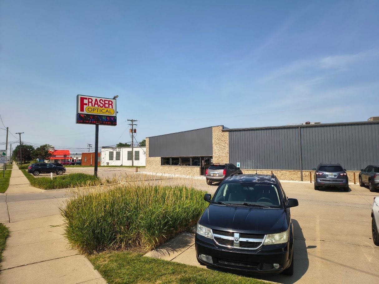 Parking lot beside a large gray building, with a black car in front and a colorful sign in the distance.