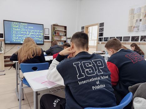 Les élèves sont assis à des pupitres dans une salle de classe, face à un tableau numérique affichant une leçon.