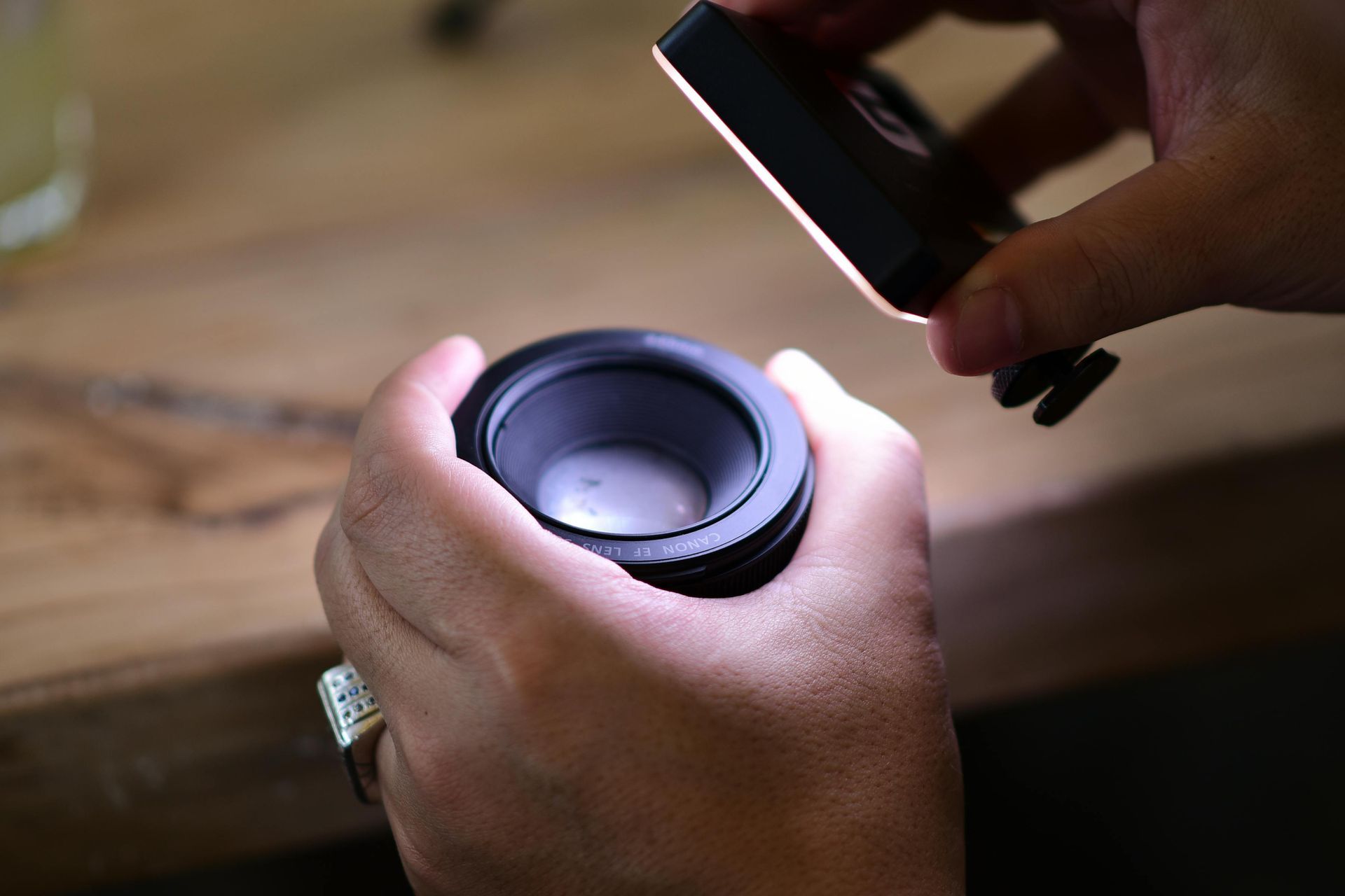 Des mains inspectent l'objectif d'un appareil photo éclairé par une petite lampe, en gros plan sur une table en bois.