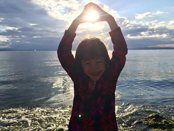 Person standing by the water with arms raised overhead, sun shining through hands at sunset