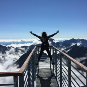 Person standing on a mountain viewpoint with arms raised above the clouds under a blue sky