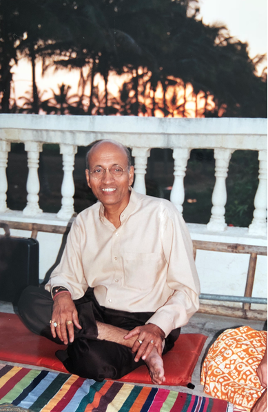 Man sitting cross-legged on a mat indoors, wearing a light shirt, smiling beside a white railing.