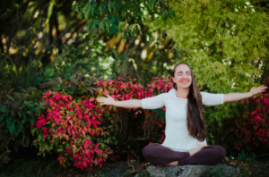 Person meditating outdoors in a garden, seated cross-legged with arms outstretched beside red and green foliage.