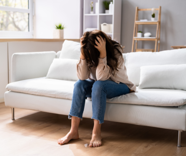 Woman sitting on a white couch with head in hands, looking distressed in a bright living room.