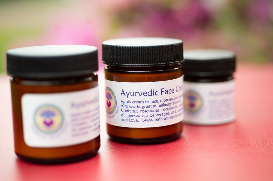 Three amber cosmetic jars with black lids on a pink surface, one labeled Ayurvedic face cream