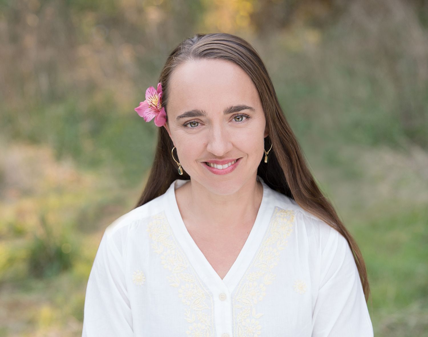 Smiling person in a white blouse with a pink flower in their hair, standing outdoors.