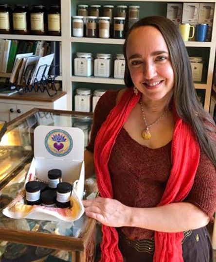 Woman in red shawl smiling, holding a tray of jars in a shop with shelves of products.
